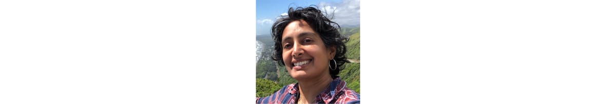 A photo of me, an Indian American woman in my early 40s, with short wavy black hair, smiling in front of distant green hills and blue sky.